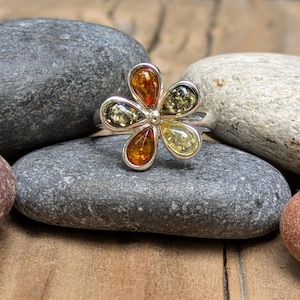 May include: A silver ring featuring a flower-shaped setting with five teardrop-shaped amber stones in varying shades of translucent orange, dark green, and pale yellow, all encased in polished silver metal frames.