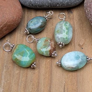 Puede incluir: Cinco colgantes de piedras preciosas pulidas de forma ovalada en tonos verde azulado, verde menta y azul claro. Cada piedra est&aacute; acentuada con tapas de metal plateado con peque&ntilde;os detalles de cuentas y una anilla circular para colgar.