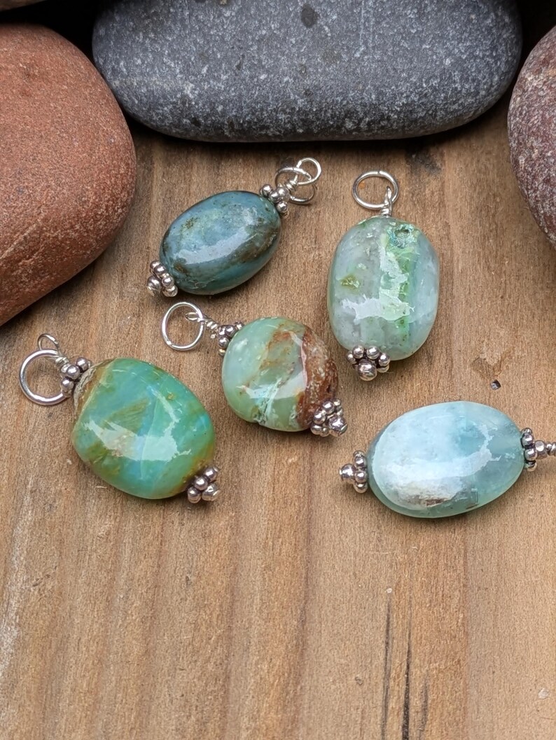 Puede incluir: Cinco colgantes de piedras preciosas pulidas de forma ovalada en tonos verde azulado, verde menta y azul claro. Cada piedra est&aacute; acentuada con tapas de metal plateado con peque&ntilde;os detalles de cuentas y una anilla circular para colgar.