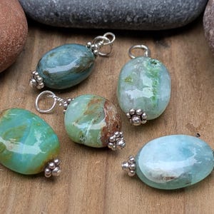 Puede incluir: Cinco colgantes de forma ovalada con piedras pulidas y multicolores en tonos verde azulado, verde claro y azul p&aacute;lido. Cada piedra est&aacute; decorada con una tapa de metal plateado y una peque&ntilde;a anilla circular para sujetarla a un collar o pulsera.