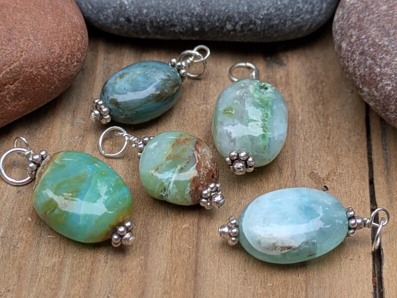 Puede incluir: Cinco colgantes de forma ovalada con piedras pulidas y multicolores en tonos verde azulado, verde claro y azul p&aacute;lido. Cada piedra est&aacute; decorada con una tapa de metal plateado y una peque&ntilde;a anilla circular para sujetarla a un collar o pulsera.