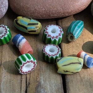 May include: A selection of vibrant glass beads in diverse shapes and patterns. The beads showcase stripes, floral motifs, and a palette of colours including green, white, red, blue, and yellow. The beads are arranged on a wooden surface.