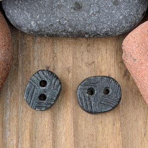 May include: Two dark gray, oval-shaped buttons with two holes each. The buttons have a textured surface with a crosshatch pattern. The buttons are on a wooden surface, with rocks in the background.
