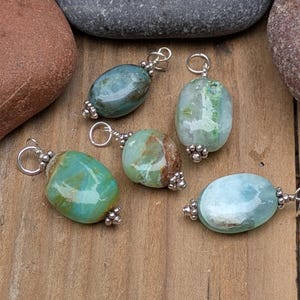 Puede incluir: Cinco colgantes de piedras preciosas pulidas de forma ovalada en tonos verde azulado, verde y azul claro. Cada piedra est&aacute; envuelta en alambre plateado con un peque&ntilde;o lazo de metal en la parte superior y cuentas plateadas decorativas en la base.