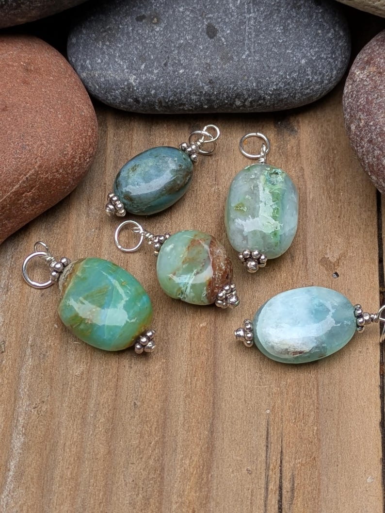 Puede incluir: Cinco colgantes de piedras preciosas pulidas de forma ovalada en tonos verde azulado, verde y azul claro. Cada piedra est&aacute; envuelta en alambre plateado con un peque&ntilde;o lazo de metal en la parte superior y cuentas plateadas decorativas en la base.