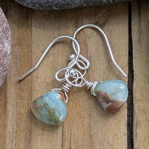 May include: A pair of handcrafted earrings featuring teardrop-shaped, blue-green gemstones with brown and red inclusions. The stones are wire-wrapped in silver-colored metal and suspended from silver-colored ear wires. The earrings are displayed on a wooden surface.