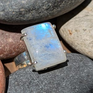 May include: A rectangular moonstone ring with a silver band. The moonstone is a translucent white with blue and iridescent flashes. The ring is set on a collection of smooth, gray and brown stones.