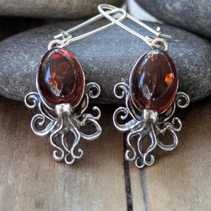 May include: A pair of silver-toned earrings featuring a dark amber-colored oval stone. The earrings are designed with an octopus motif, with the stone serving as the head and the tentacles forming the lower portion of the design.