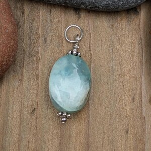 Puede incluir: Un colgante que presenta una piedra preciosa de color azul claro, lisa y de forma ovalada, con inclusiones blancas. La piedra est&aacute; decorada con peque&ntilde;as cuentas de metal plateado pulido en la parte superior e inferior, e incluye una anilla plateada.