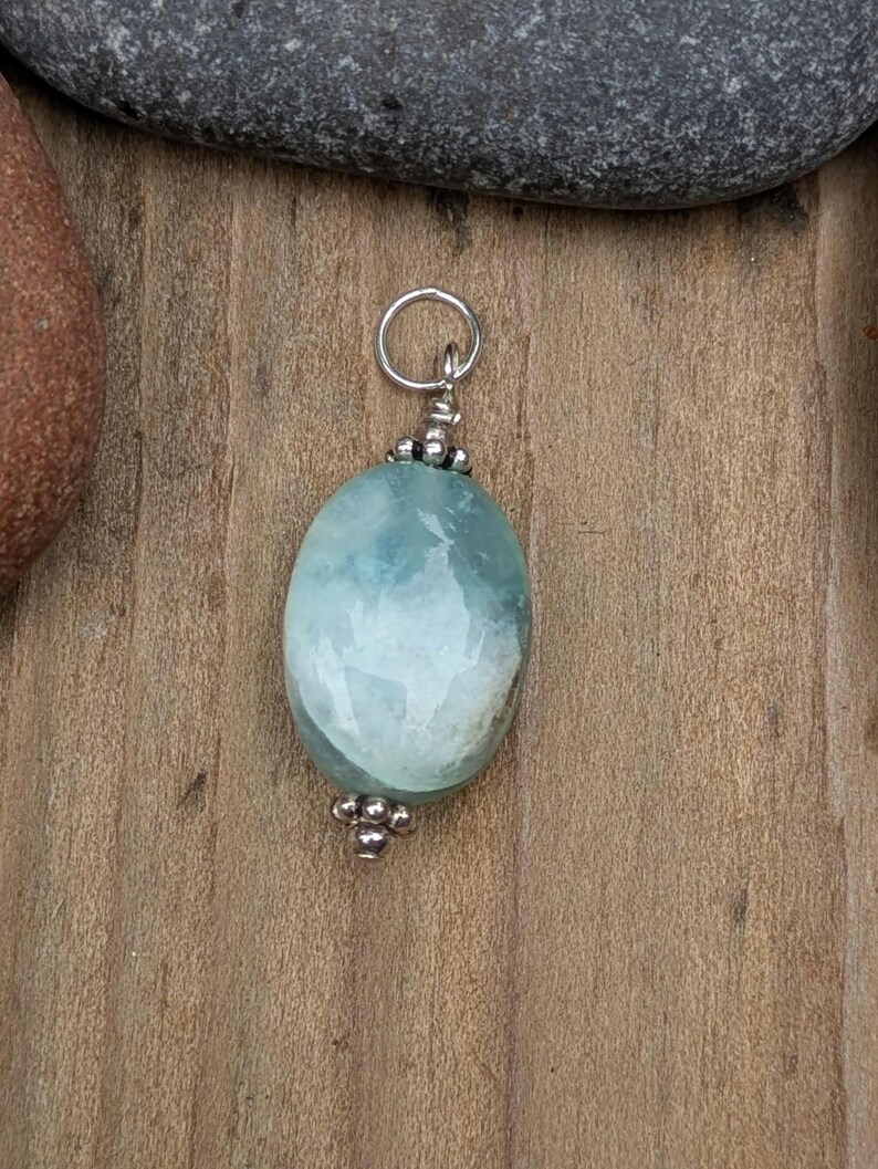 Puede incluir: Un colgante que presenta una piedra preciosa de color azul claro, lisa y de forma ovalada, con inclusiones blancas. La piedra est&aacute; decorada con peque&ntilde;as cuentas de metal plateado pulido en la parte superior e inferior, e incluye una anilla plateada.