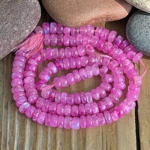 Puede incluir: Un collar de cuentas de piedras preciosas rosas enrollado sobre una superficie de madera. Las cuentas son de un color magenta vibrante, con una calidad ligeramente translúcida. Una borla rosa está unida. Las cuentas tienen aproximadamente 0,5 cm de diámetro.
