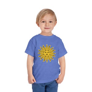 May include: A blue toddler t-shirt featuring a yellow sun design with a smiling face. The sun has pointed rays and a black outline. The shirt is made of a soft material and is suitable for children.