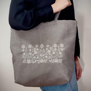 May include: Grey tote bag featuring a white floral pattern. The bag is made from a textured material and has a rectangular shape. The floral design, comprising various flowers and leaves, is centrally placed on the front.