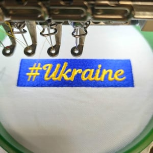 May include: A close-up of an embroidery machine stitching a blue patch with the text "#Ukraine" in yellow thread.