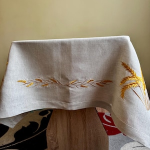 Puede incluir: Un mantel de mesa de lino gris claro con espigas de trigo bordadas en oro. El mantel está drapeado sobre una mesa de madera y está sobre una alfombra estampada.