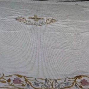 Pode incluir: Um tecido branco com uma borda floral bordada em ouro, rosa e azul. O design inclui um emblema central com uma cruz. O tecido parece ser uma toalha de mesa ou um pano de altar, com um padrão detalhado e ornamentado.