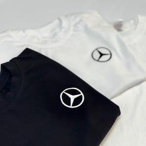 Może przedstawiać: Wyświetlane są dwie koszulki z krótkim rękawem, jedna czarna i jedna biała. Każda koszulka ma mały, haftowany emblemat na piersi. Emblemat to trójramienna gwiazda w okręgu. Koszulki są złożone i ułożone w stos.