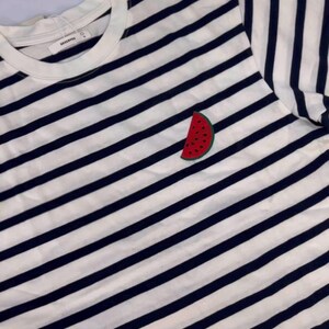 May include: White and navy blue striped t-shirt with a watermelon patch. The t-shirt has a round neckline and short sleeves. The watermelon patch is red with black seeds and a green rind.