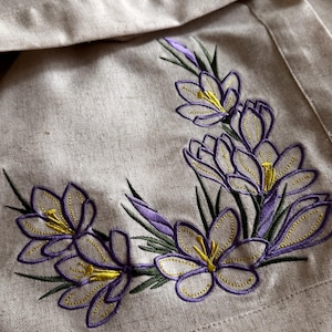 May include: Beige linen fabric with embroidered purple and yellow crocus flowers. The floral design features detailed stitching, with green leaves and stems. The fabric is folded, showing the embroidered flowers in the corner.