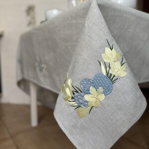 May include: A grey linen tablecloth with embroidered yellow flowers and blue Easter eggs. The tablecloth is draped over a white table.