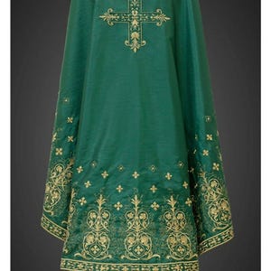 Puede incluir: Vestidura litúrgica verde esmeralda con cruz y motivos florales bordados en oro. La vestidura presenta detalles intrincados en los bordes y una prenda interior blanca que asoma por la parte inferior. El diseño es simétrico y formal.