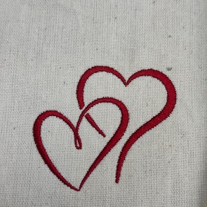 May include: Two red hearts intertwined, embroidered on a white fabric background.