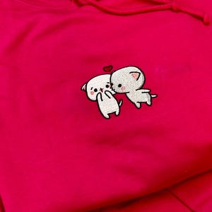 Può includere: Una felpa con cappuccio rosa acceso con coulisse. La felpa presenta un disegno ricamato di due gatti dei cartoni animati che si baciano, con un piccolo cuore rosso sopra. Il disegno è bianco con contorni neri.