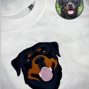 May include: White t-shirt with an embroidered Rottweiler dog design. The dog has black fur, brown accents, and a pink tongue. A circular image of a Rottweiler is in the upper right corner. The shirt has an XXS label.