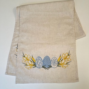 以下が含まれることがあります： ベージュのリネン製のテーブルランナーで、青と白のイースターエッグと黄色の花が刺繍されています。
