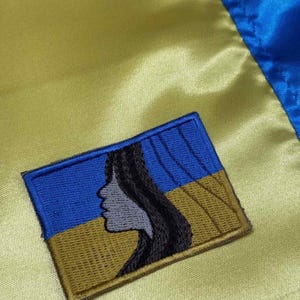 May include: A blue and gold embroidered patch with a stylized silhouette of a woman's face. The patch is on a yellow fabric background.