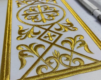 May include: Close-up of a white fabric with an intricate gold embroidered design. The embroidery features a cross within a circle, geometric shapes, and floral patterns. The gold thread creates a textured, ornate appearance, suggesting a religious or ceremonial item.