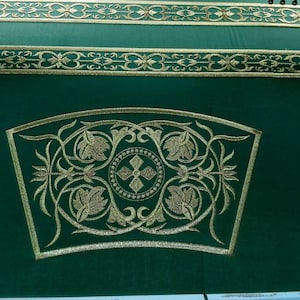 May include: Close-up of a green textile with gold embroidery. The design features floral and geometric patterns, including a central cross within a circle. Decorative gold bands run across the top.