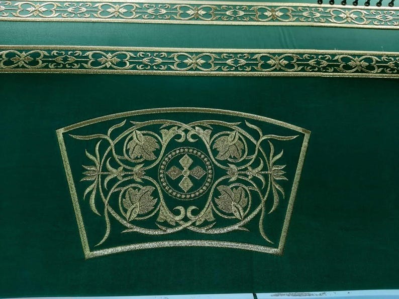 May include: Close-up of a green textile with gold embroidery. The design features floral and geometric patterns, including a central cross within a circle. Decorative gold bands run across the top.