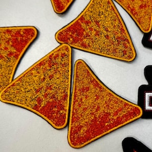 May include: Close-up of several embroidered patches in a triangular shape, resembling a tortilla chip. The patches have a yellow and red color scheme with a black border. The patches are arranged on a light gray surface.