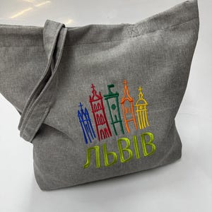 May include: Gray tote bag with embroidered colorful buildings and the word "ЛЬВІВ" in yellow-green. The bag has a simple design with a single handle and is made of a textured fabric.
