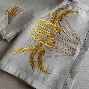 Puede incluir: Dos servilletas de lino grises con espigas de trigo bordadas en oro. Las servilletas están dobladas y superpuestas.