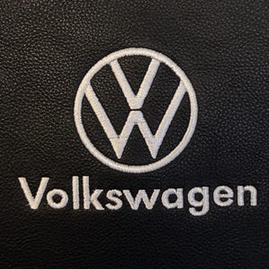 Puede incluir: Un material negro texturizado con el logotipo de Volkswagen bordado en blanco, que consiste en un emblema circular con una V y una W estilizadas en su interior, seguido de la palabra Volkswagen en una fuente sans-serif debajo del icono.