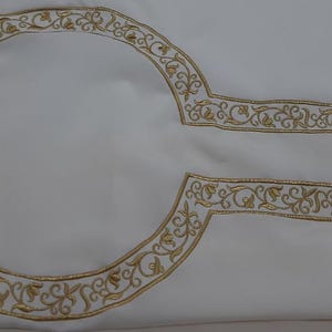 May include: Ivory fabric with gold embroidered floral designs. The embroidery forms curved and rectangular shapes, likely part of a garment or textile art piece. The detailed stitching adds a touch of elegance.