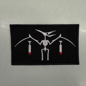 May include: Black rectangular patch featuring a white pterodactyl skeleton with outstretched wings. Two white, stylized bombs with red tips are positioned on either side of the pterodactyl. The patch is likely an embroidered emblem.