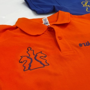 May include: Two polo shirts, one orange and one blue, are displayed. The orange shirt features a blue embroidered chess-themed design and the text "#uk". The blue shirt is visible in the background.