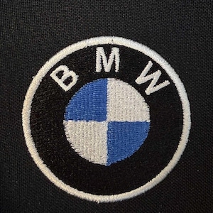 Op de afbeelding: Een ronde geborduurde patch met het BMW-logo, voorzien van de witte letters B, M en W op een zwarte achtergrond. Het midden is verdeeld in vier kwadranten met afwisselend blauwe en witte kleuren, omringd door een witte cirkelvormige rand.