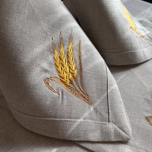 Op de afbeelding: Twee grijze linnen servetten met geborduurde tarwehalmen in geel en bruin. De servetten zijn gevouwen in driehoeken en liggen op een grijs linnen tafelkleed.