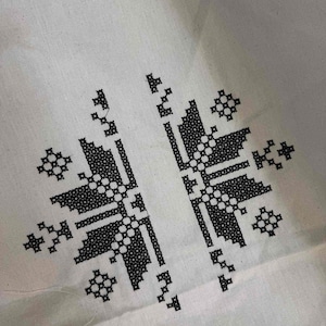 May include: Close-up of white fabric with a black cross-stitch pattern. The design features symmetrical floral and geometric shapes. The fabric has a slightly textured appearance, and the stitching is detailed.