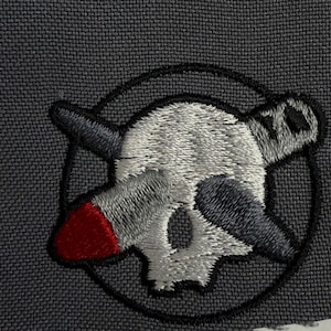 May include: Embroidered patch featuring a skull with crossed horns and a missile. The skull is white with black accents, and the missile has a red tip. The patch is set against a dark gray fabric background.