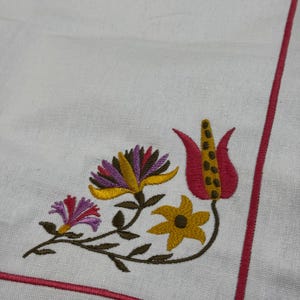 Könnte beinhalten: Weißer Stoff mit gesticktem Blumendesign. Die Stickerei zeigt eine Ansammlung von Blumen in Gelb-, Lila- und Rottönen mit grünen Stielen und Blättern. Ein rosa Rand umrahmt den Stoff.