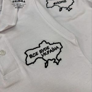May include: White polo shirts with black embroidered outlines of the Ukraine map. One shirt has the text "ВСЕ БУДЕ УКРАЇНА" and the other has "#HOME". The shirts have a collared neckline and button closure.