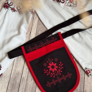 Könnte beinhalten: Eine schwarz-rote bestickte Tasche mit einem langen schwarzen Riemen. Die Tasche hat ein rotes Blumendesign und geometrische Muster. Der Hintergrund umfasst ein weißes besticktes Hemd und eine Holzoberfläche.