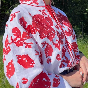 Könnte beinhalten: Weiße Bluse mit roter Blumenstickerei. Die Bluse hat lange Ärmel und einen hohen Kragen, mit rotem Besatz und einer dekorativen Kordel. Die Stickerei ist detailliert und bedeckt die Ärmel und die Vorderseite.