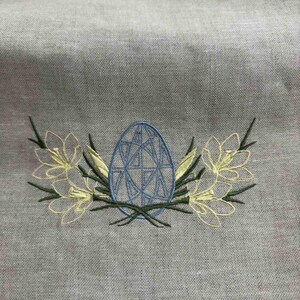 May include: Embroidered design on a light gray fabric featuring a blue egg with a geometric pattern surrounded by yellow flowers and green stems.