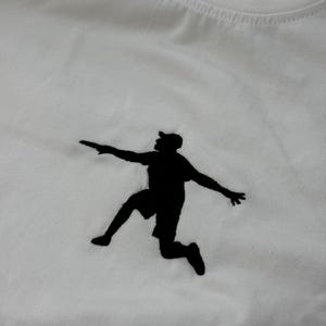 May include: White t-shirt featuring a black silhouette of a person jumping with arms outstretched. The design is centered on the chest. The shirt is made of a soft, white fabric.